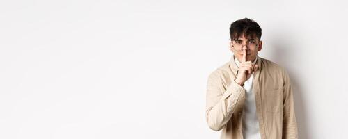 Cheerful handsome man in glasses share a secret, making surprise, lean at camera with hush gesture and gentle smile, telling to be quiet, show taboo sign, white background photo