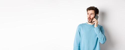Technology concept. Intrigued young man looking left impressed, talking on mobile phone, calling someone, standing over white background photo
