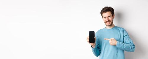 E-commerce and shopping concept. Smiling caucasian man pointing finger at empty smartphone screen, showing online offer, white background photo