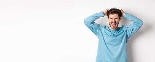 Mad young man pull hair oyt of head and screaming angry, shouting frustrated, standing hateful against white background photo