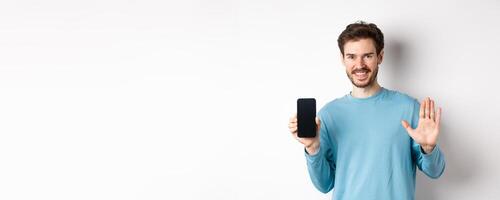 Handsome caucasian man with beard, showing empty smartphone screen and number five, raising hand to wave and say hello, standing on white background photo