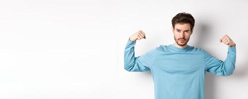 Confident and strong macho man flexing biceps, showing strength in muscles after gym workout, standing over white background photo