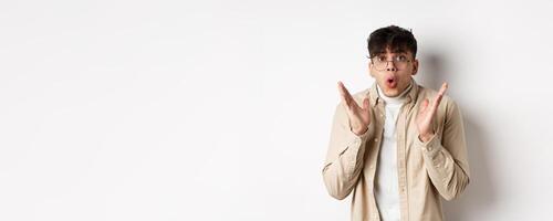Portrait of surprised hipster guy in glasses checking out awesome promo, say wow and gasping amazed, raising hands up with disbelief, standing on white background photo