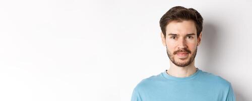 Close up of young handsome man with beard, smiling at camera with confidence, standing over white background photo
