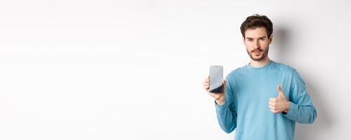 Young man showing empty smartphone screen and thumb up, recommend mobile application or online shopping offer, standing over white background photo