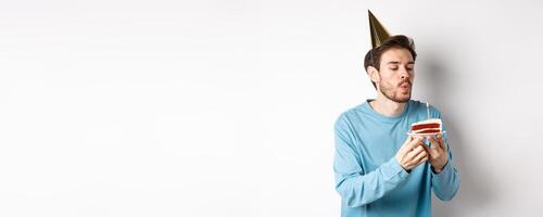 Celebration and holidays concept. Happy silly guy blowing candle on cake and making wish, standing in party hat over white background photo