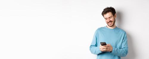 Portrait of modern caucasian man with beard, wearing casual clothes, reading smartphone screen and smiling, networking via mobile phone, standing over white background photo
