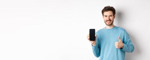 Smiling young man showing empty smartphone screen and thumbs up, praising good application, recommend mobile app, standing over white background photo