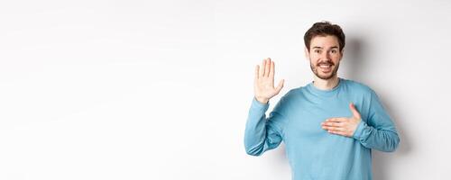 Smiling hosent man raising arm and hold hand on heart, making promise tell truth, swear or give oath, standing over white background photo