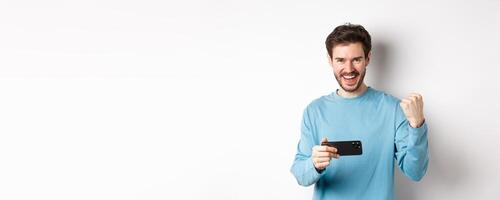 Lucky man winning in mobile game, holding smartphone and saying yes, triumphing with fist pump, standing over white background photo