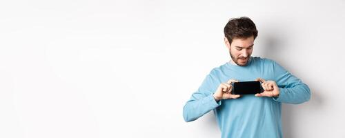 E-commerce and shopping concept. Caucasian man showing blank mobile screen and looking at it, standing curious against white background photo
