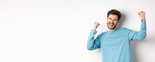 Image of happy young man triumphing, feeling like champion, winning and screaming in cheer, standing over white background photo