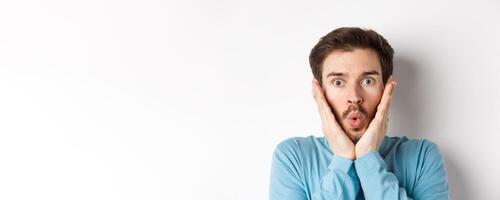 Close-up of impressed man checking out special offer, saying wow and touching face with amazed expression, stare at camera in awe, standing over white background photo