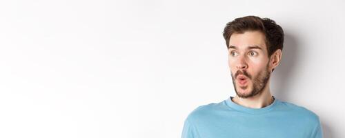Close-up of impressed caucasian man saying wow, looking left amazed, checking out promotion deal, white background photo