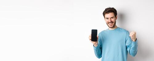 Image of happy man showing empty smartphone screen and celebrating, smiling with rejoice and fist pump, achieve online goal, standing over white background photo