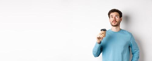 Image of young man drinking takeaway coffee and looking up with thoughtful face, tasting new flavour, standing over white background photo