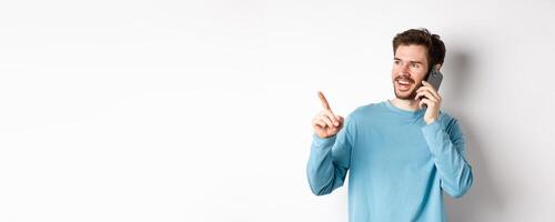 Technology concept. Young man talking on mobile phone and pointing left, having call on smartphone, standing over white background photo