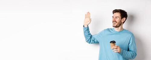 Friendly young man drinking coffee from takeaway cup and saying hello, waving hand left and smiling, standing over white background photo