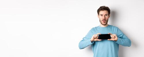 E-commerce and shopping concept. Image of impressed man saying wow and showing blank mobile phone screen in horizontal, standing over white background photo