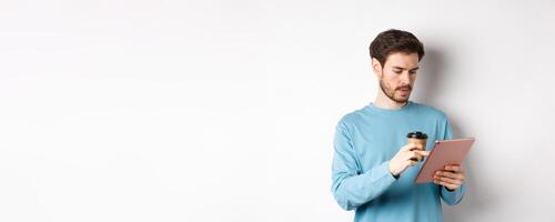 Man browsing online in digital tablet and drinking coffee from paper cup, standing over white background photo