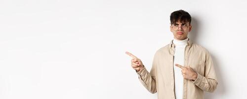 Disappointed guy in glasses looking and pointing left, sulking displeased, complaining on bad offer, standing upset on white background photo