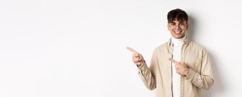Portrait of handsome caucasian young man in glasses showing logo, smiling and pointing fingers left, inviting to check out promo deal, white background photo
