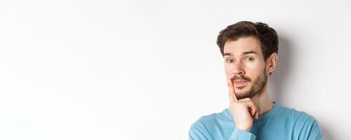 Image of thoughtful young man with beard, making choice, touching lip and looking pensive at camera, deciding something, white background photo