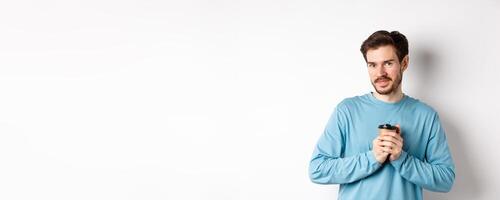 Handsome caucasian man warm-up hands with cup of coffee, smiling at camera and looking tenderly, standing on white background photo