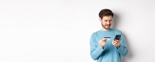 E-commerce and shopping concept. Smiling man order online, copy credit card number to mobile phone app, standing over white background photo