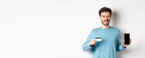Young man in casual shirt showing empty smartphone screen and plastic credit card, winking and smiling at camera, standing on white background photo