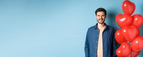 Cute boyfriend looking happy and smiling, standing near Valentines day heart balloons on blue background photo