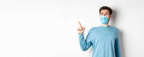 Coronavirus, health and quarantine concept. Surprised guy looking in awe and pointing at upper left corner, standing in medical mask and sweatshirt over white background photo