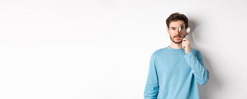 Man searching for something, looking through magnifying glass at camera, standing on white background photo