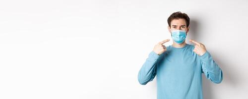 Coronavirus, health and quarantine concept. Young caucasian man in casual clothes pointing at his medical mask, asking to use preventive measures during pandemic, white background photo
