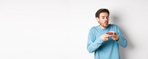 Celebration and holidays concept. Happy young man blowing candle on birthday cake, making wish on bday, standing over white background photo