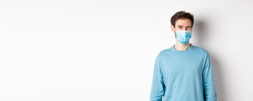 Covid-19, pandemic and social distancing concept. Cheerful caucasian man in face mask winking at camera, looking happy, standing over white background photo