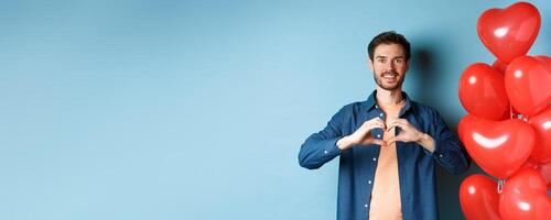 Happy valentines day. Boyfriend in love showing heart gesture to lover and smiling, standing near red balloons on blue background photo