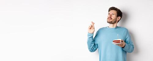 Celebration and holidays concept. Handsome young man celebrating birthday, making wish with bday cake in hand, standing over white background photo