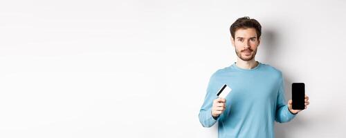 Handsome caucasian man showing empty smartphone screen and plastic credit card, standing over white background photo