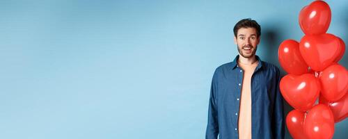 Happy valentines day. Excited smiling guy standing near red hearts balloons and looking at camera, blue background photo
