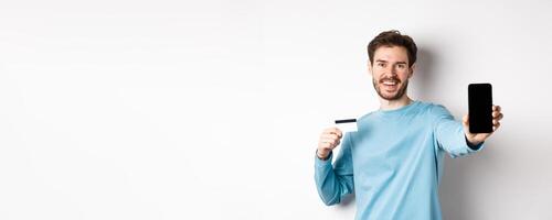 E-commerce and shopping concept. Smiling caucasian man showing plastic credit card and empty smartphone screen, recommending online app, white background photo