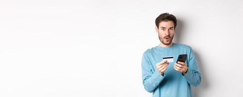 E-commerce and shopping concept. Excited man looking amused while making order online, holding plastic credit card and smartphone, standing on white background photo