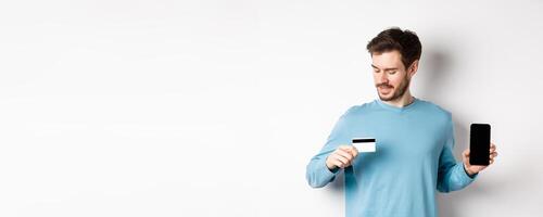 Handsome young man with beard, showing empty smartphone screen and looking at plastic credit card, standing over white background photo