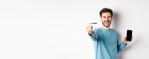 E-commerce and shopping concept. Happy man showing plastic credit card and screen of smartphone, recommending banking app, standing on white background photo