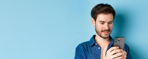 Close up portrait of happy young man reading mobile sreen, networking in smartphone, standing on blue background photo