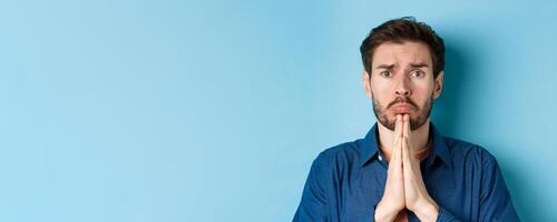 Sad young man begging for help, apoligizing and sobbing miserable, standing on blue background photo