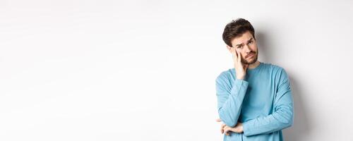Sad young man looking frustrated, touching temple of head and frowning uneasy, standing against white background photo