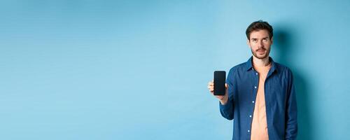 Annoyed young guy showing empty smartphone screen and roll eyes up bothered, standing on blue background photo