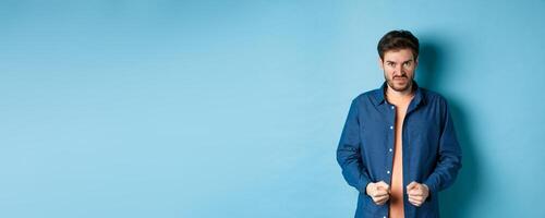 Angry and pissed-off man clench fists and stare with disdain at camera, trying to keep himself together, look with rage, standing on blue background photo