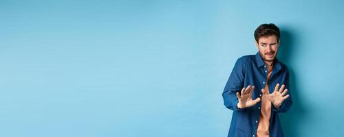 Man cringe and stay away from something nasty, looking aside and stretch out hands to reject bad deal, standing on blue background photo